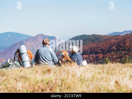 Father and son travelers with their beagle dog sit together in mountain valley with beautiful hills view Stock Photo