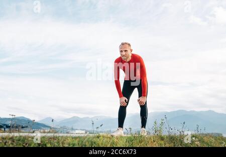 The wide angle shoot of a Man dressed in red long sleeve shirt very tired after the jog  by the road with mountain background and resting to lean forw Stock Photo