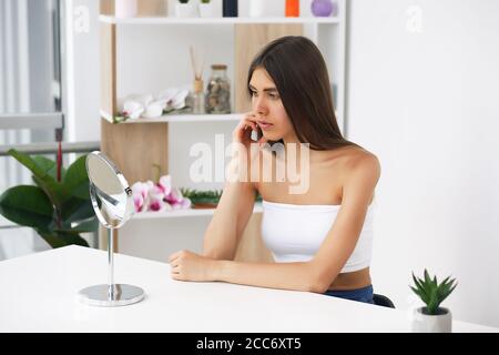 Young brunette girl takes care of her skin, standing in front of a mirror, enjoying beauty treatments for herself, smiling tenderly Stock Photo