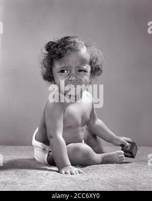 1940s 1950s UNHAPPY 1 YEAR OLD AFRICAN-AMERICAN BABY GIRL WEARING DIAPER SITTING PLAYING WITH TOY BLOCK GRIMACING LOOKING AWAY  - n314 HAR001 HARS BLOCK FACIAL INFANT WORRY LIFESTYLE FEMALES MOODY HOME LIFE COPY SPACE FULL-LENGTH AWAY DIAPER EXPRESSIONS TROUBLED B&W CONCERNED SADNESS AFRICAN-AMERICANS AFRICAN-AMERICAN BLACK ETHNICITY MOOD CONCEPTUAL GLUM GRIMACING JUVENILES MISERABLE BABY GIRL BLACK AND WHITE HAR001 OLD FASHIONED AFRICAN AMERICANS Stock Photo