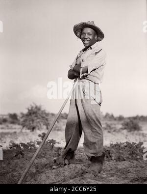 1930s SMILING AFRICAN-AMERICAN MAN TOBACCO FARMER WEARING STRAW HAT STANDING LEANING ON HOE LOOKING AT CAMERA NORTH CAROLINA USA - n68 HAR001 HARS 1 FACIAL LAUGH PLEASED JOY LIFESTYLE SATISFACTION JOBS POOR RURAL UNITED STATES COPY SPACE FULL-LENGTH PERSONS UNITED STATES OF AMERICA FARMING MALES CONFIDENCE EXPRESSIONS AGRICULTURE B&W NORTH AMERICA EYE CONTACT NORTH AMERICAN SKILL OCCUPATION HAPPINESS SKILLS CHEERFUL STRENGTH TOBACCO AFRICAN-AMERICANS AFRICAN-AMERICAN FARMERS LOW ANGLE BLACK ETHNICITY LABOR PRIDE ON EMPLOYMENT OCCUPATIONS CONCEPTUAL HANDLE JOYFUL HOE SHARECROPPER EMPLOYEE Stock Photo