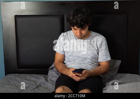 Young Hispanic man with long hair, resting on a bed, using a phone during quarantine Stock Photo