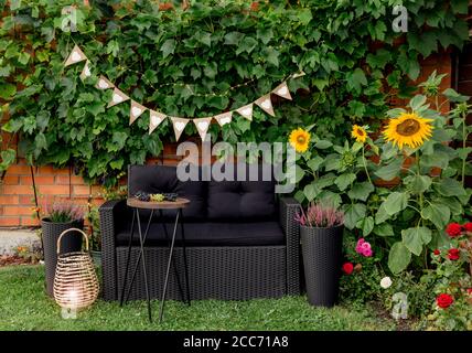 Black modern polyrotang garden furniture chair, metal wire side table. Green grape vines plants growing on red brick wall as decorative element. Bamb Stock Photo