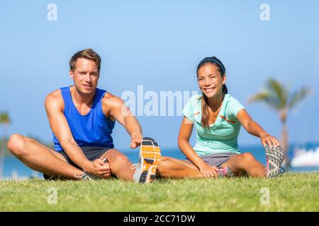 Stretching happy runners group doing leg stretch together on run class outside in summer grass doing jogging warm-up. Interracial young couple, Asian Stock Photo