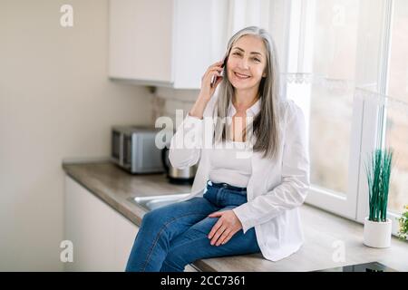 retired people and technologies. Pleasant cheerful senior gray haired woman in white shirt and denim, talking on cell phone, while sitting on the Stock Photo