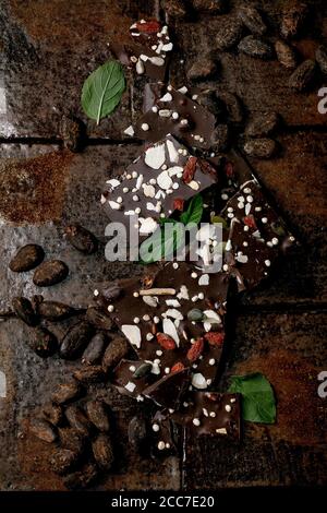 Handmade chopped dark chocolate with different superfood additives seeds and goji berries, with cocoa beans and fresh mint over dark ceramic tile back Stock Photo