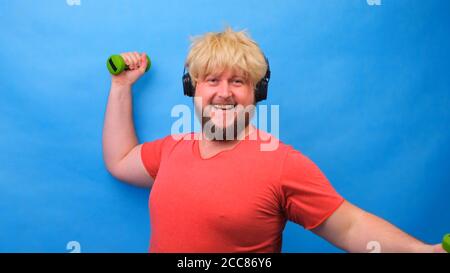 Funny fat man with dumbbells doing exercises on a blue background. Stock Photo