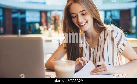 Happy caucasian girl with red hair and freckles working in a cafeteria with a laptop and papers while drinking a coffee Stock Photo