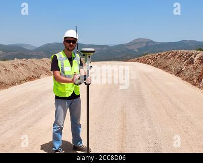 Geodetic engineer surveyor in white hard hat doing measurements with GNSS satellite receiver during road construction works. Stock Photo