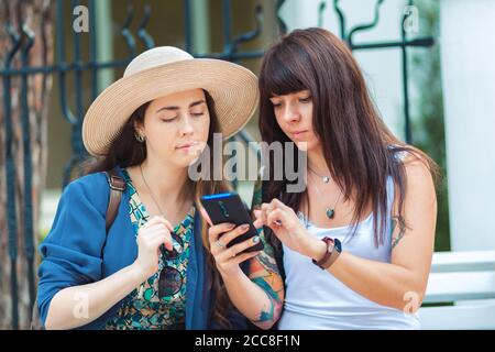 Two women sit on the street, and look at their smartphone, making purchases in an online store. One woman holds a Bank card in her hand. The concept o Stock Photo