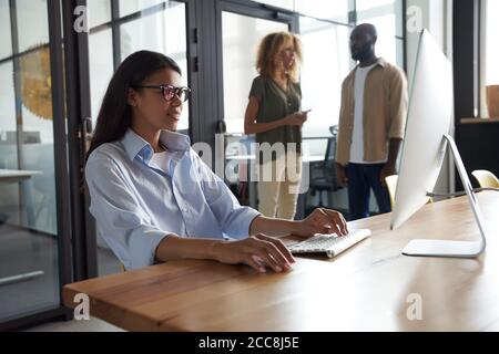 Young mixed race woman, female office worker working on computer while sitting at her workplace in the modern office, colleagues standing on the Stock Photo