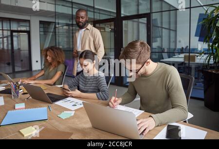 Successful business people. Group of young multiracial people, coworkers working together in modern office, using laptops, smartphone, making some Stock Photo