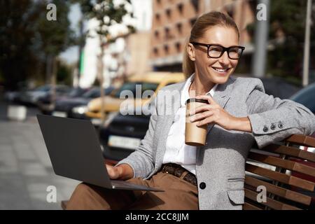 Working from everywhere. Portrait of a happy beautiful business woman drinking coffee and using laptop while sitting on the bench outdoors Stock Photo