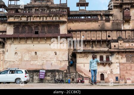 Orchha, Madhya Pradesh, India - March 2019: A young Indian man wearing sunglasses walking with the beautiful exterior facade of the ancient Jehangir M Stock Photo