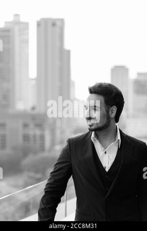 Thoughtful happy Indian businessman smiling while enjoying the view at rooftop against view of the city Stock Photo