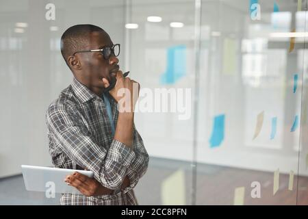 Pensive African businessman in eyeglasses standing in front of glass wall and looking at adhesive notes on it he thinking about new ideas Stock Photo