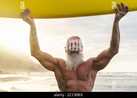 Happy fit senior having fun surfing at sunset time - Sporty bearded man training with surfboard on the beach
