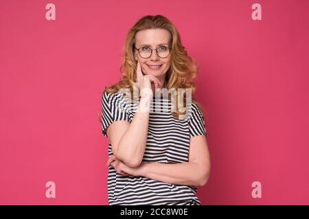 Mature woman in glasses smiling at camera standing against pink background Stock Photo