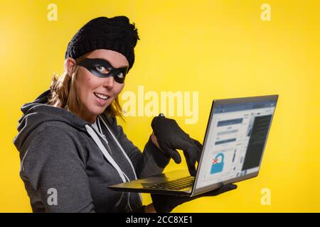 The concept of cybercrime and hacking. Portrait of woman in black hat, gloves and mask holding laptop with browser window about password lock. Yellow Stock Photo