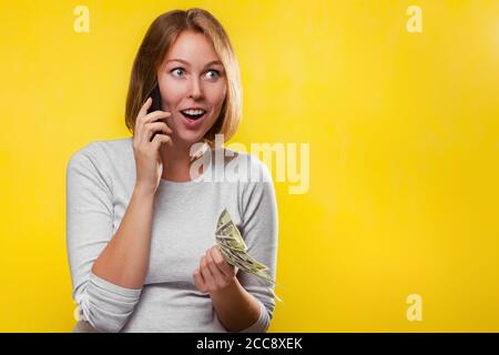 Finance and Bank cards. A young blonde holds a wad of dollars in her hands, and with surprise calls on her mobile phone. Yellow background and copy sp Stock Photo