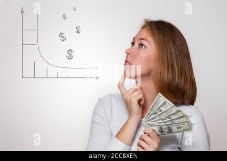 Finance and banking. A young blonde holds a wad of dollars in her hands and looks thoughtfully at the drawn diagram. White background. Stock Photo