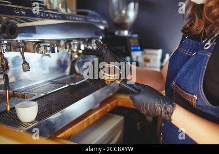 Barista wearing medical latex black gloves,preparing coffee drink with espresso machine in coffee shop cafe.Woman presses ground coffee using tamper Stock Photo