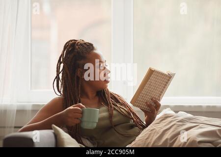 African young woman lying on sofa drinking coffee and reading an interesting book at home Stock Photo