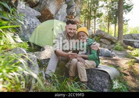 Father and son sitting on stone and using mobile phone outdoors during hiking Stock Photo