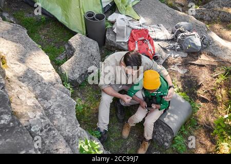 High angle view of father embracing his son while they sitting on stone and watching something on mobile phone during their trip Stock Photo