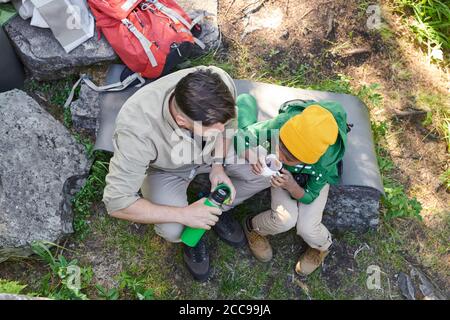 High angle view of father and his son sitting on stone and drinking tea from vacuum flask during their camping Stock Photo