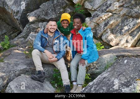 Portrait of happy family of tourists sitting on rocks outdoors and smiling at camera during camping Stock Photo