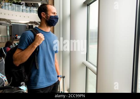 young man wearing protective face mask, traveling in the time of covid19 pandemic inside an airport, looking out of window with smiling and positive e Stock Photo