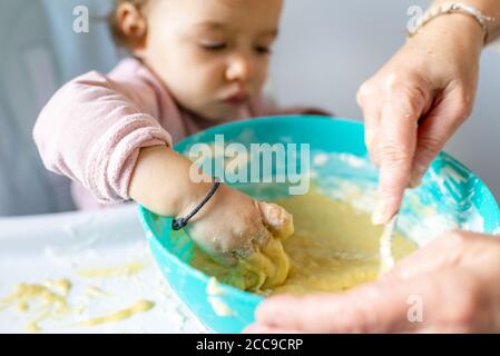 Little girl (16 months) sitting in her high chair making a cake with her mother. Here, reaching the cake batter in a green plastic container. Reproduc