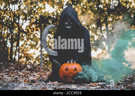 Girl with halloween make up, holding a sickle with blood and a pumpkin with a smoke bomb inside - Holidays, party, scary and autumn concept - Focus on Stock Photo
