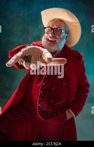 Choosing you. Modern stylish Santa Claus in red fashionable suit and cowboy's hat on dark background. Looks like a rockstar. New Year and Christmas eve, celebration, holidays, winter's mood, fashion. Stock Photo