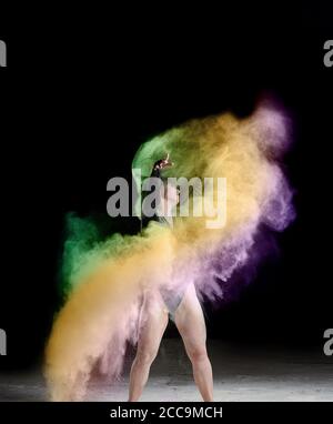 beautiful caucasian woman in a black bodysuit with a sports figure is dancing in a multicolored cloud of flour on a black background Stock Photo