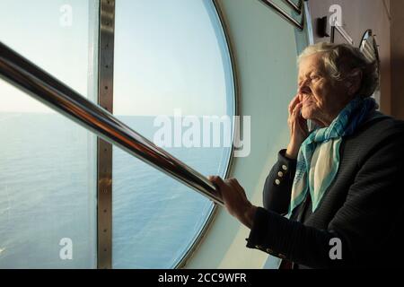 A kind old woman looks thoughtfully at the sea through the porthole of the ferry. Concept: solitude, recollection Caring for lonely pensioners. High quality photo Stock Photo