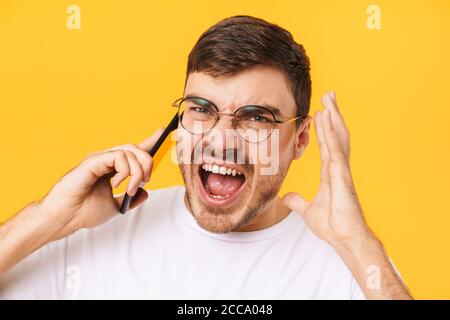 Photo of angry caucasian man in eyeglasses talking on smartphone and screaming isolated over yellow background Stock Photo