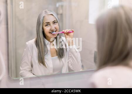 Pretty retired woman with long gray hair, wearing white shirt, brushing her teeth in the bathroom at home, standing in front of big mirror. Reflection Stock Photo