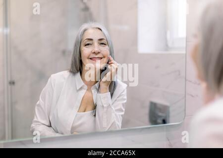 Beautiful retired gray haired lady standing in bathroom in front of the mirror, looking at camera with smile, while applying anti-aging face cream and Stock Photo