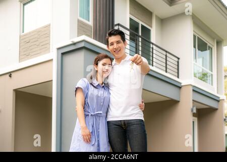 Portrait of Asian young couple standing and hugging together and holding house key looking happy in front of their new house to start new life. Family Stock Photo