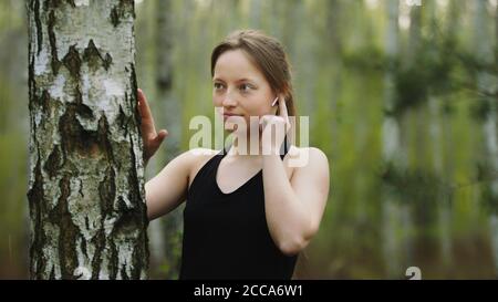 Young caucasian woman in the nature. Listening the music on the wireless headphones and touching the tree bark. Slow motion. High quality photo Stock Photo