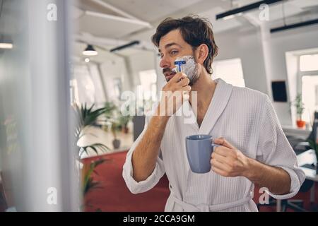 Handsome young man in bathrobe shaving at work Stock Photo