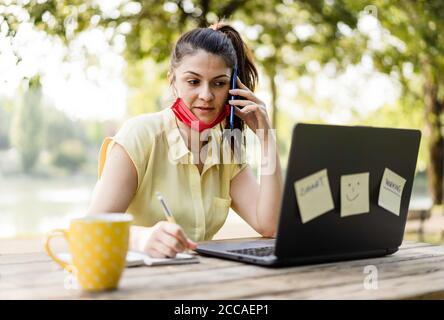 Young woman wearing lowered protective face mask using laptop computer outdoors - Female freelancer or manager working on laptop at park - Remote work Stock Photo