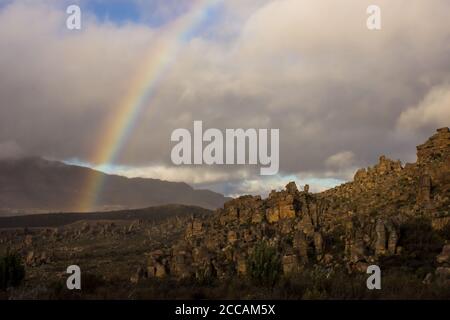 A rainbow in the Cederberg Mountains of South Africa with the weathered rock formations of the Window rocks in the foreground, lit by the last of the Stock Photo