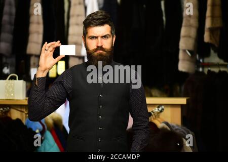 Customer with beard and business card. Shop assistant with empty paper. Finance and shopping concept. Man with strict face holds card on furry coats racks background, defocused Stock Photo