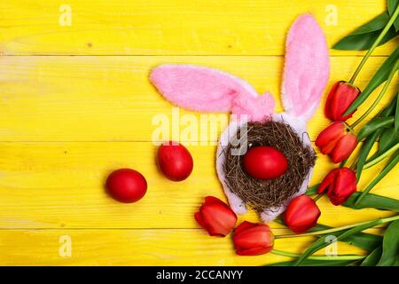 Bunch of flowers near pink Easter eggs in hay nest with fluffy bunny ears on. Spring holiday composition. Tulips in pink or red colors on yellow wooden background. Happy holiday concept. Stock Photo
