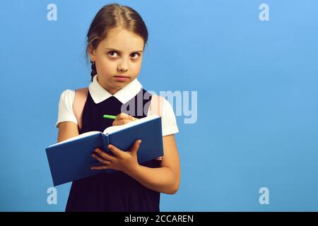 School girl with thoughtful face isolated on blue background. Back to school and education concept. Girl writes in big blue notebook with green pen. Pupil in school uniform with braid and backpack Stock Photo