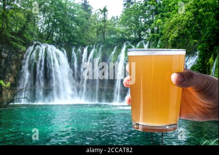 Man holding glass of light beer on big waterfall background. Relax, rest among nature. Stock Photo