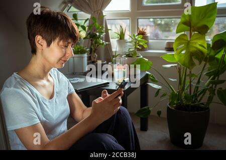 Close up mixed race woman in casual home clothes serfing in her smart phone sitting near coffee table with closed laptop and many green potted plants Stock Photo